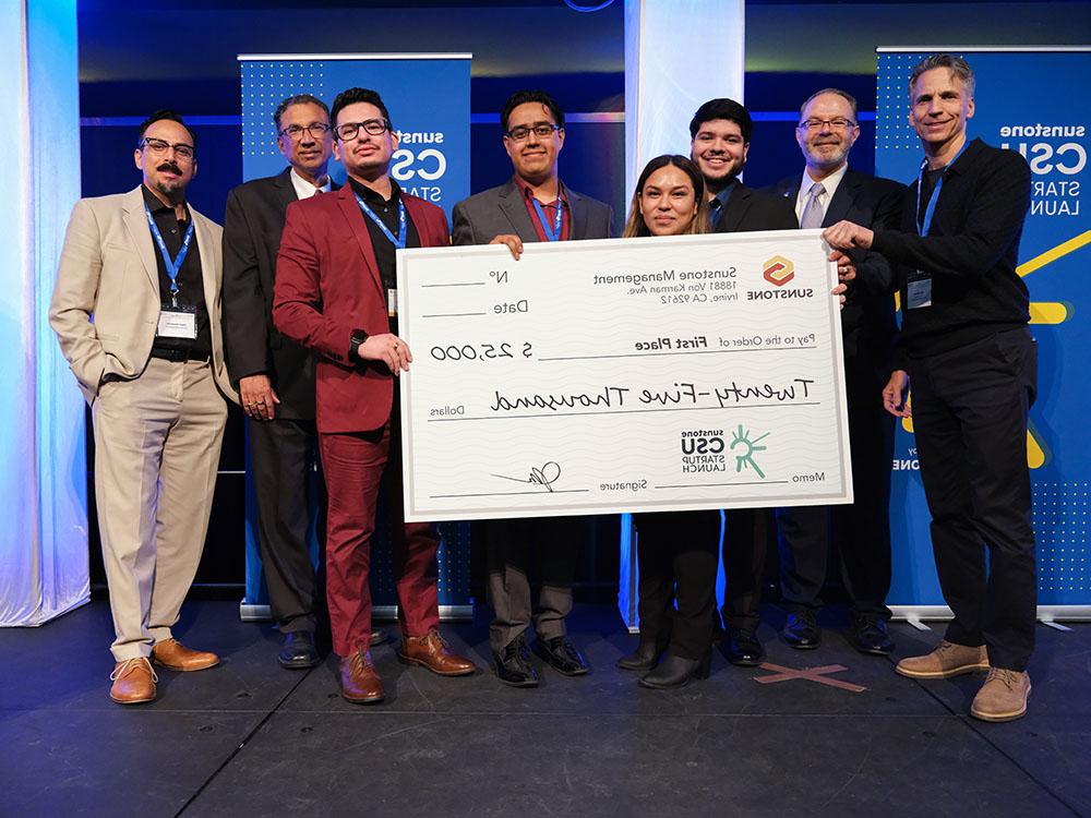 Four college-age startup founders pose onstage with two sponsors, two hosts, and a giant check for $25,000