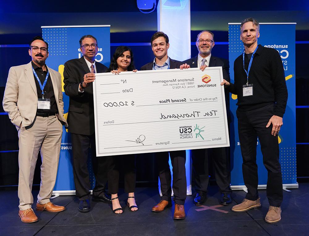 Two college-age startup founders pose onstage with two sponsors, two hosts, and a giant check for $10,000