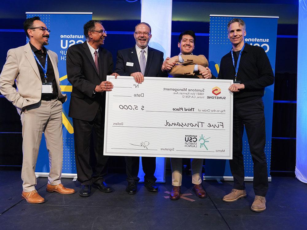 A college-age startup founder poses onstage with two sponsors, two hosts, and a giant check for $5,000