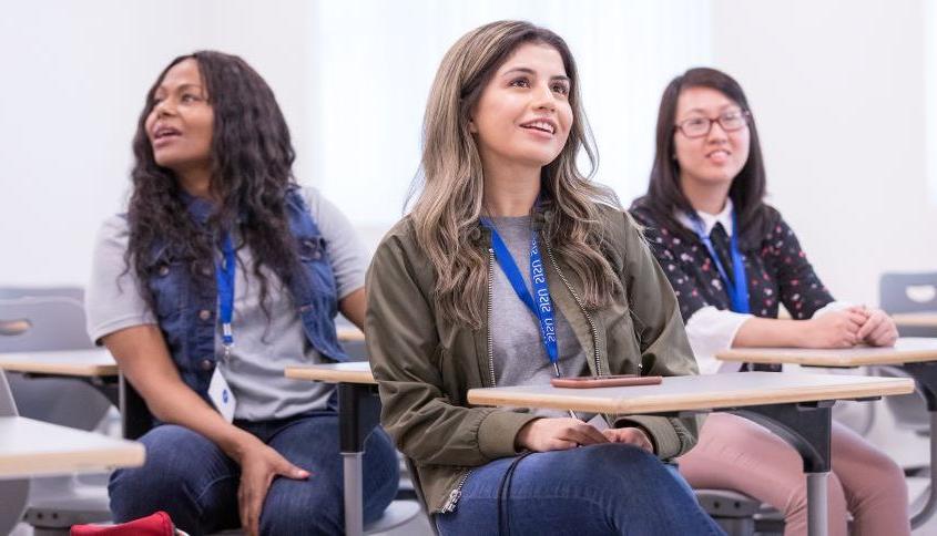 Three students smile while listening at desks in a presentation setting